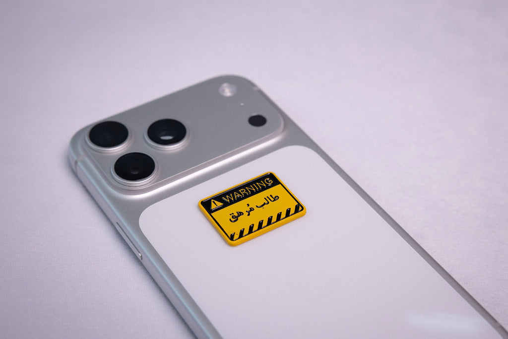 Yellow-and-black WARNING sign sticker reading طالب مُرهق (exhausted student), applied to a phone case near the camera bump