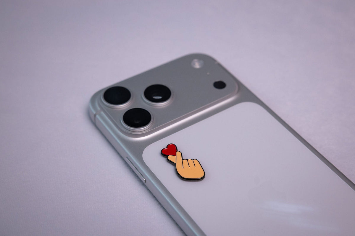 Classic K-pop-style finger heart gesture (thumb and index finger crossed with a small red heart), applied to a phone case near the camera bump