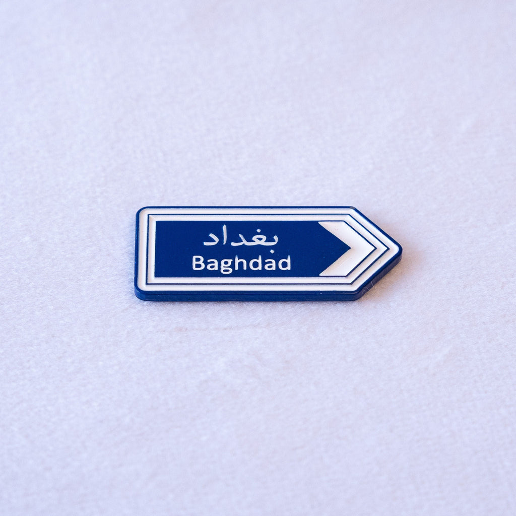 A blue highway sign cut as an arrow, بغداد above Baghdad, with a white chevron pointing the way.