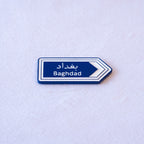 A blue highway sign cut as an arrow, بغداد above Baghdad, with a white chevron pointing the way.