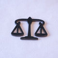 Balanced scales of justice die-cut in matte black metal, the pans cut through as open triangles, as a metal sticker.