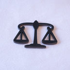 Balanced scales of justice die-cut in matte black metal, the pans cut through as open triangles, as a metal sticker.
