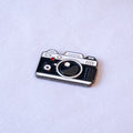 A retro film SLR in black and silver with a big round lens, a chrome top plate, and a small red shutter button, as a metal sticker.