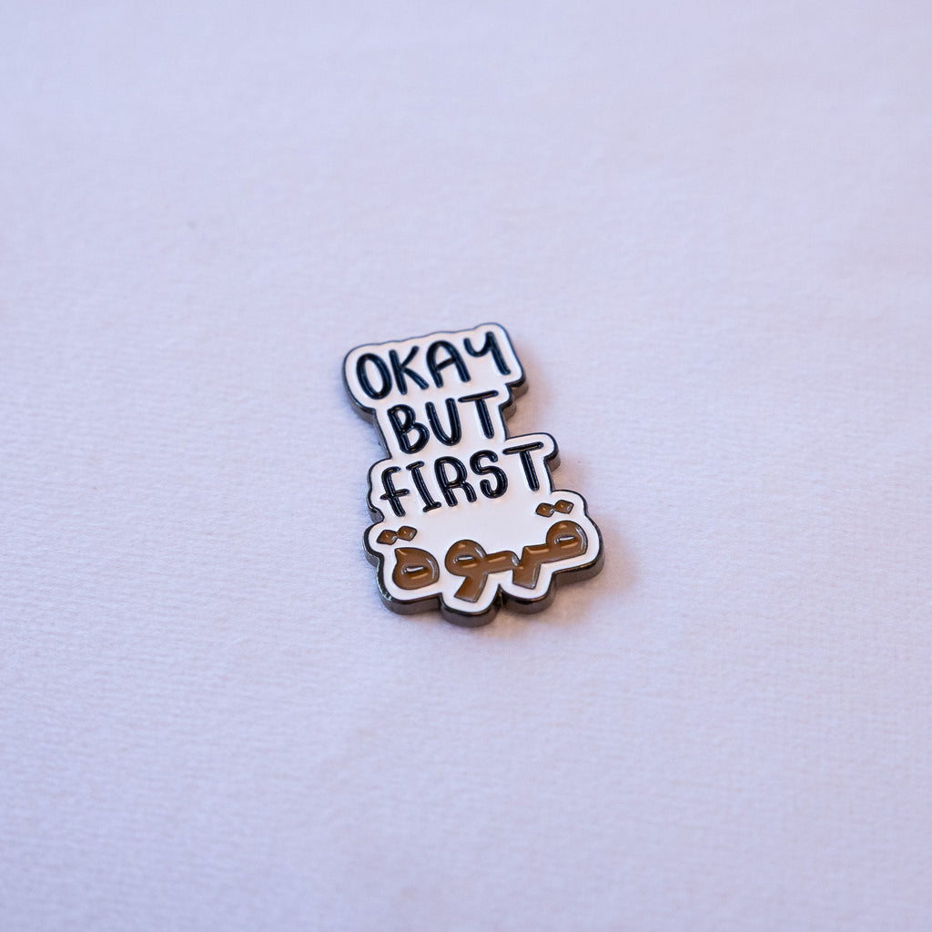 OKAY BUT FIRST in navy hand-lettered capitals with قهوة beneath it in warm brown, as a metal sticker.
